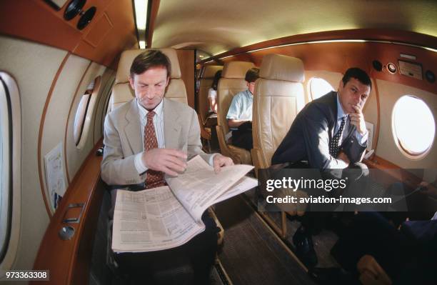 Businessmen and women sitting in the passenger cabin of a Dassault Falcon 20, one man reading a newspaper with another looking more tense thinking...