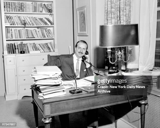 Producer David Merrick conducts business as he sits behind his desk which is loaded with manuscripts.