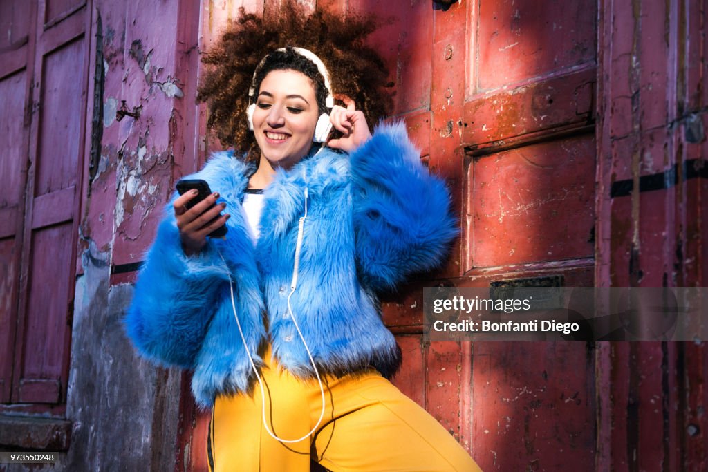 Portrait of woman with afro hair wearing fur coat, listening to music through headphones on smartphone