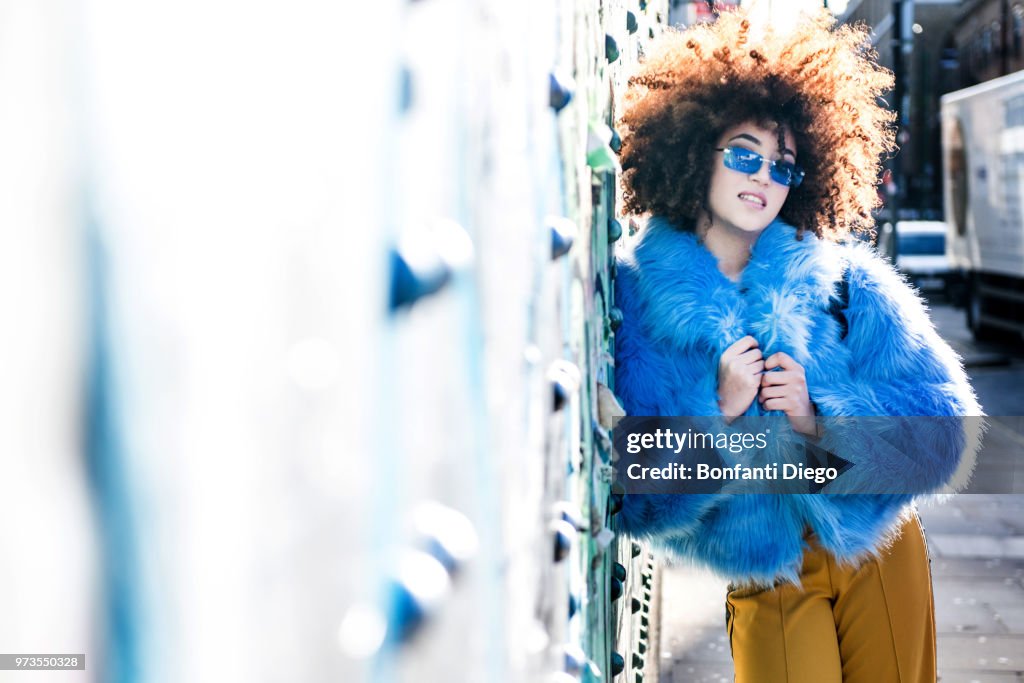 Portrait of woman with afro hair wearing fur coat, leaning against wall looking at camera