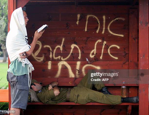 Jewish settler prays as a soldier sleeps in a bus stop where a graffiti reads: "My brother, don't throw me away" 17 August 2005 in the Gaza Strip...