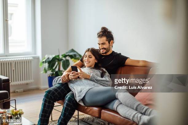 young couple with mobile phone relaxing on sofa - due persone foto e immagini stock