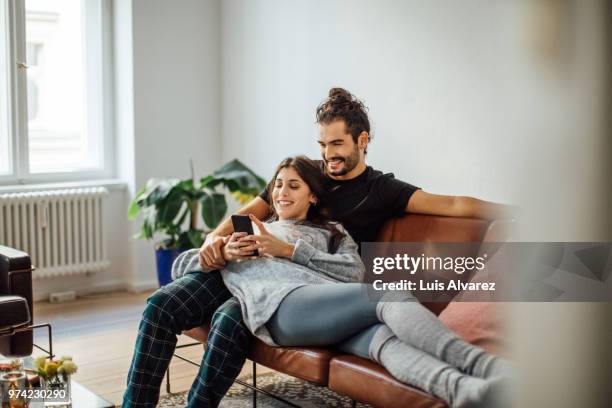 young couple with mobile phone relaxing on sofa - woman couple at home stockfoto's en -beelden