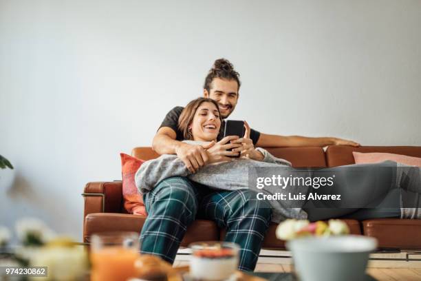 young couple with smart phone relaxing on sofa - couple smartphone stockfoto's en -beelden
