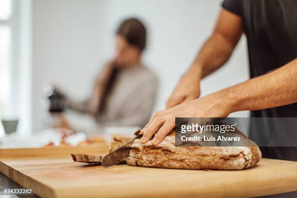 young man slicing bread on cutting board in kitchen - loaf of bread stock-fotos und bilder