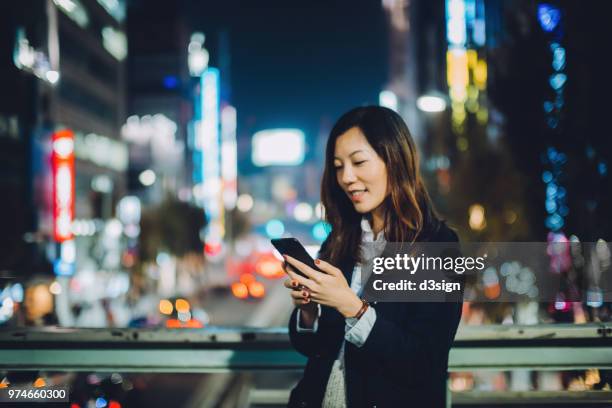 young asian woman requesting a ride with smartphone in downtown city street at night - smart street light stock pictures, royalty-free photos & images