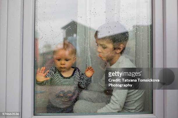 two sad children, baby boy and older brother, sitting in front f the window while raining - casa famiglia foto e immagini stock