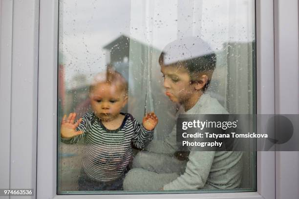 two sad children, baby boy and older brother, sitting in front f the window while raining - weeshuis stockfoto's en -beelden