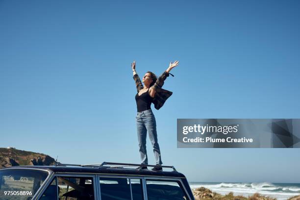 young woman on roof of car with arms outstretched - free imagens e fotografias de stock