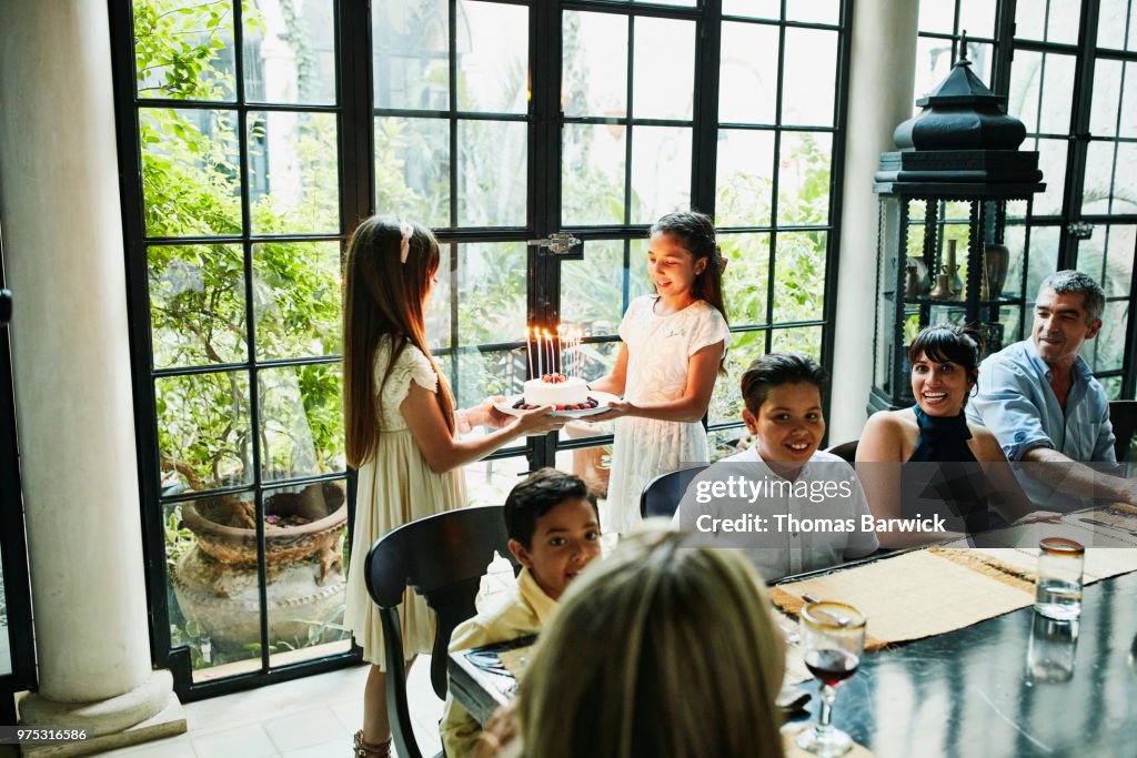 Female cousins bringing birthday cake with candles to dinner table during family birthday party