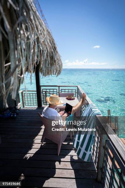 business retreat man sat outside relaxing and working with feet up on a wooden deck of a tropical overwater bungalow working remotely and wirelessly on computer and smartphone - sattel bildbanksfoton och bilder