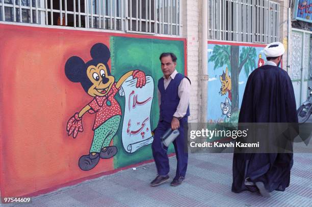 Cleric passes by a Mickey Mouse cartoon painted on the wall of a kindergarten in a street in central Qom, Iran, 3rd February 1998. Qom is a world...