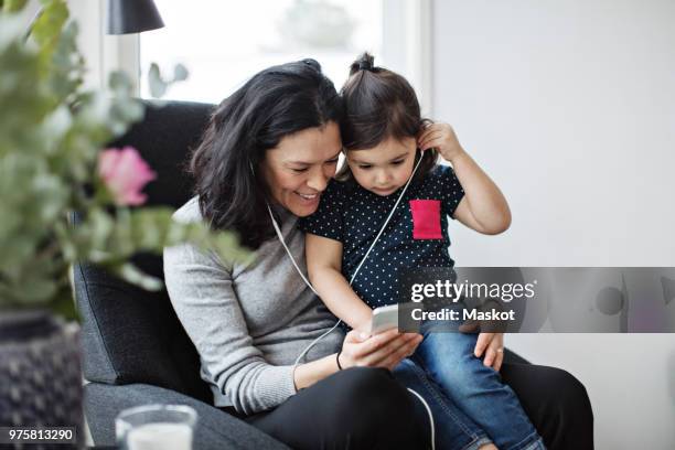 mother and daughter wearing headphones while listening music through mobile phone in living room - music room stockfoto's en -beelden