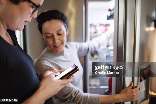 woman showing mobile phone while girlfriend opening refrigerator in kitchen - smart kitchen fotografías e imágenes de stock