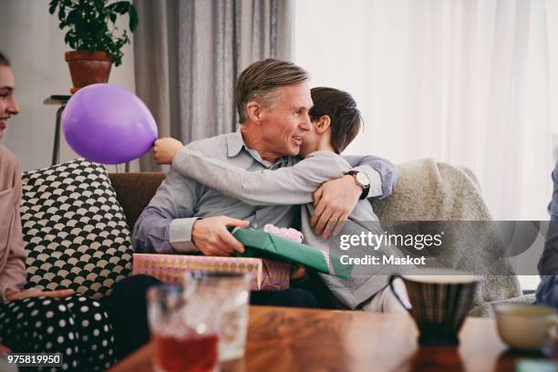 grandfather holding birthday gift while embracing grandson on sofa at home - boys birthday stock pictures, royalty-free photos & images