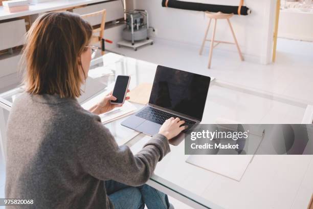 rear view of female owner using laptop and mobile phone at desk in workshop - femme de dos smartphone photos et images de collection