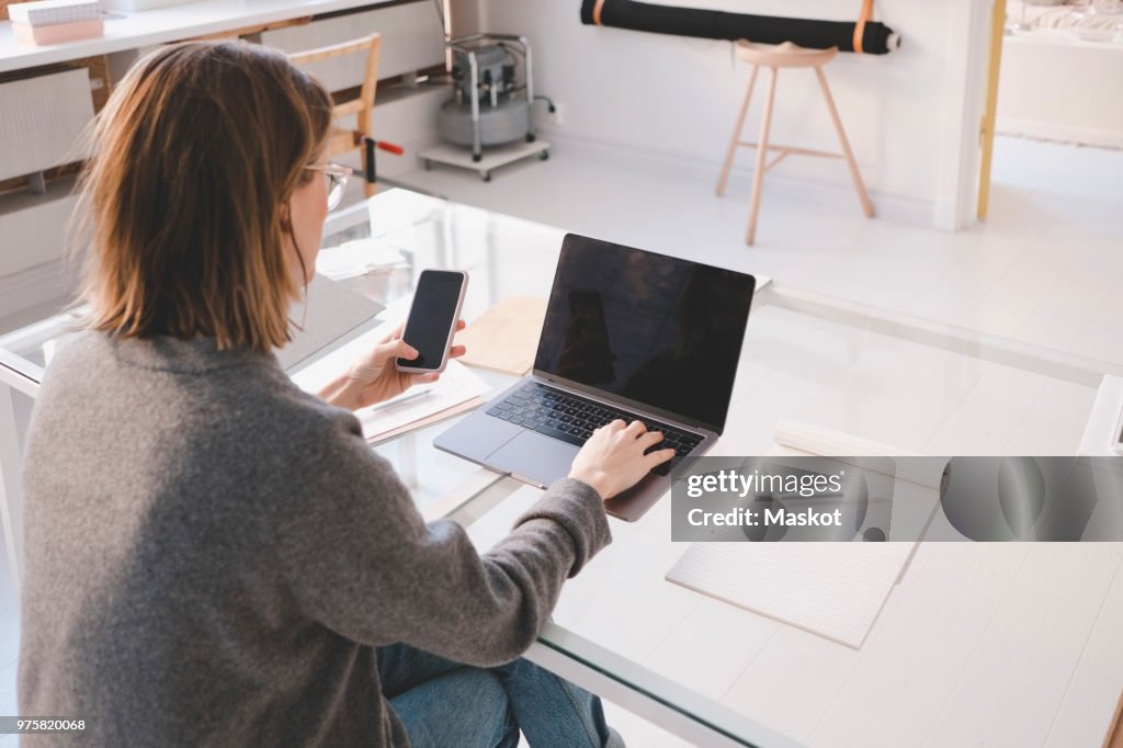 Rear view of female owner using laptop and mobile phone at desk in workshop