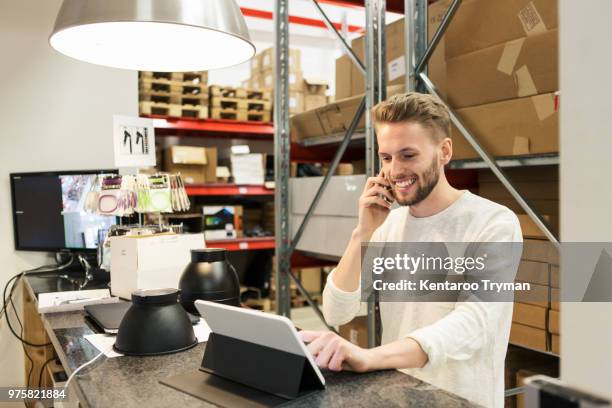 smiling man talking on mobile phone while using digital tablet in warehouse - part time job stockfoto's en -beelden