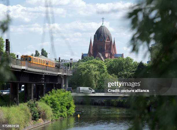 May 2018, Germany, Berlin: A U-Bahn train drives over the Landwehr Canal in the direction of the station Warschauer Strasse. The Holy Cross Church...