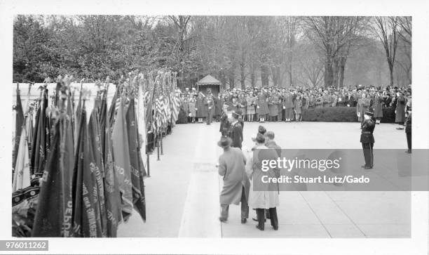 Black and white photograph, showing several photographers, a line of uniformed military personnel, and civilian spectators ringing an open, paved...