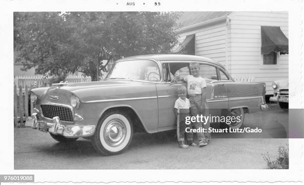 Black and white photograph, showing two small, smiling, blonde boys, likely brothers, standing in front of the driver's side door of a two-tone,...