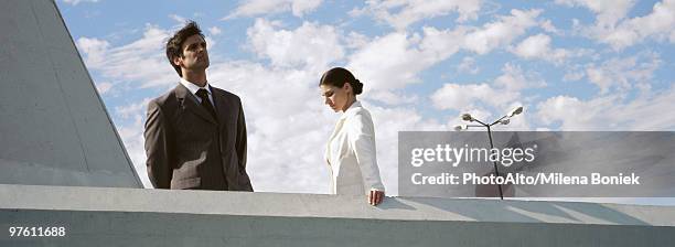 businessman and businesswoman standing on rooftop, man looking at view, woman looking down - sims stock-fotos und bilder