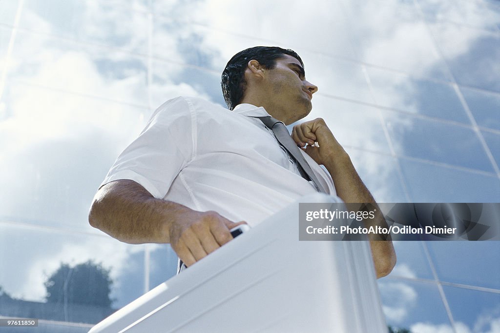 Businessman standing outside office building, carrying briefcase and adjusting collar, low angle view