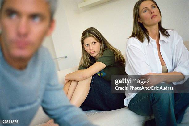 teenage girl sitting on bed hugging knees, parents sitting nearby looking away with air of frustration - problemas de comunicação imagens e fotografias de stock