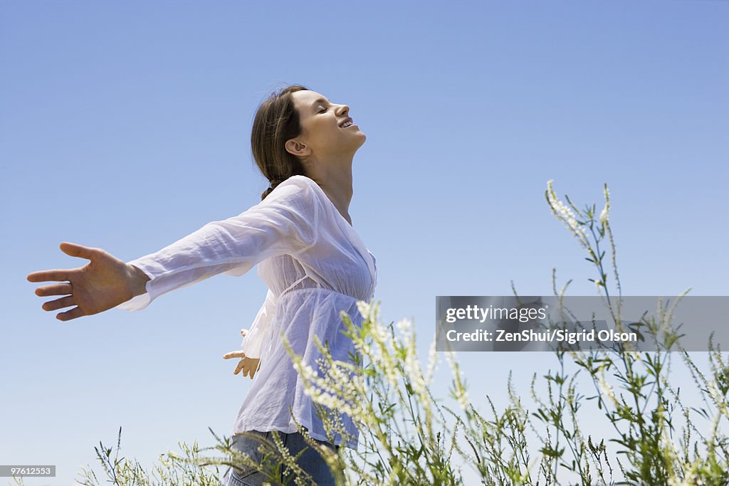 Young woman standing in tall grass with arms out, eyes closed