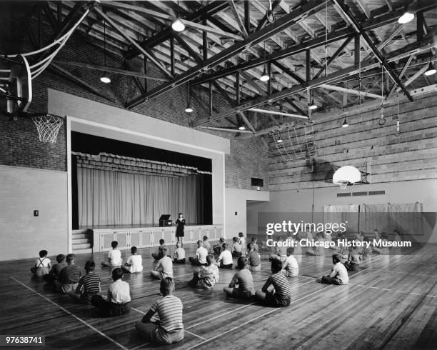 View of children seated on the floor of a grade school gymnasium as a teacher stands next to a stage, Oak Ridge, Tennessee, July 12, 1944. The city...