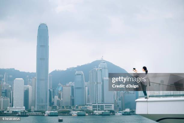 asian female traveller photographing the iconic cityscape with mobile phone from balcony - hong kong harbour stock pictures, royalty-free photos & images