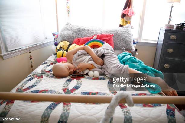 a young girl with cancer, lays on her bed looking a little nervous,  surrounded by pillows and stuffed animals. - childhood cancer stock pictures, royalty-free photos & images