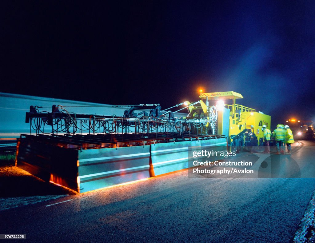 Road surfacing in progress with an asphalt spreader laying heated tarmac at night.
