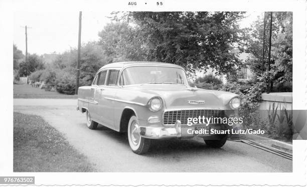 Black and white photograph, showing a two-tone, hard-top Chevrolet Bel Air, with an Ohio license plate, parked in a driveway, with grass, trees, and...