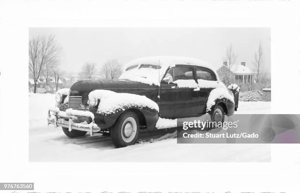 Black and white photograph, showing a three-quarter profile view of a dark, vintage, Chevrolet sedan car, parked outside in a wintery landscape, with...