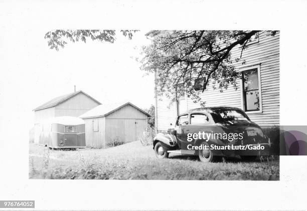 Black and white photograph, showing a three-quarter, partial profile view of a shiny, dark, vintage Chevrolet sedan, parked outside in front of a...