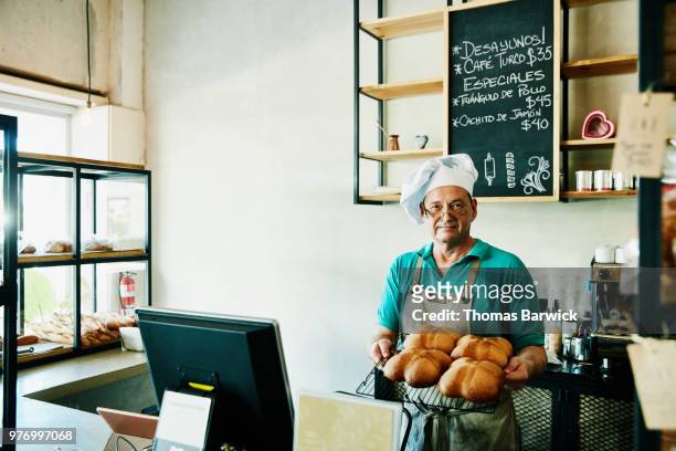 Baker standing behind counter in shop holding tray of freshly baked bread