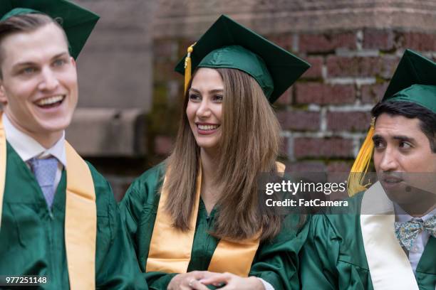 un groupe multiethnique d’étudiants s’asseoir autour et rions ensemble dans leurs robes de graduation et casquettes - sasseoir photos et images de collection