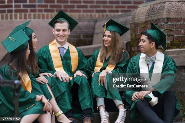 un groupe multiethnique d’étudiants s’asseoir autour et rions ensemble dans leurs robes de graduation et casquettes - sasseoir photos et images de collection