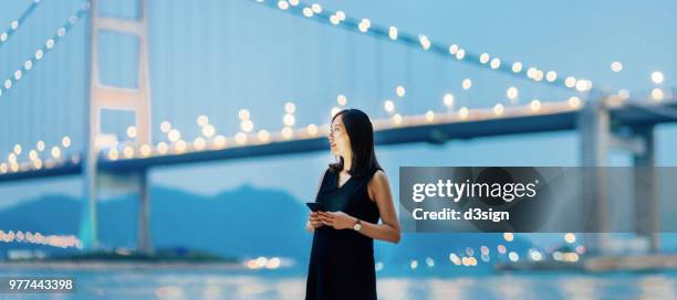 young woman using smartphone by the promenade in city, standing against modern bridge at dusk - tsing ma bridge stock-fotos und bilder