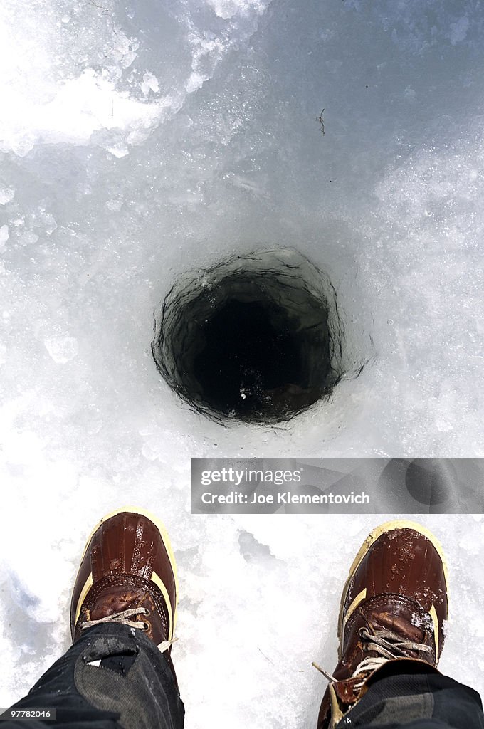 Ice fisherman's boots looking down into a dark hole in the ice.