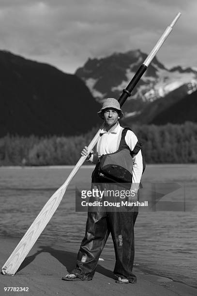 a man stands on the shores of a river for a portrait with an oar, wool hat and rain slicker overalls - rain hat stock pictures, royalty-free photos & images