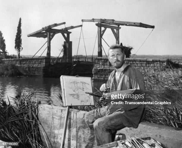 American actor Kirk Douglas sits on a river bank with a painting in a scene from 'Lust for Life,' directed by Vincent Minelli and George Cukor,...