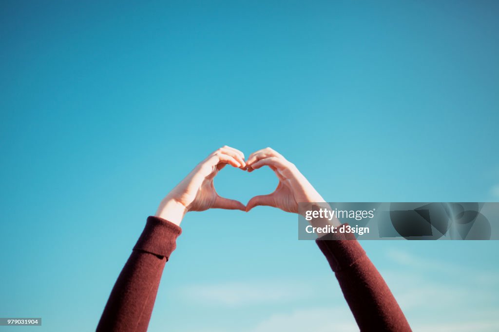 Woman's hand gesturing a heart shape over clear blue sky and warm sunlight