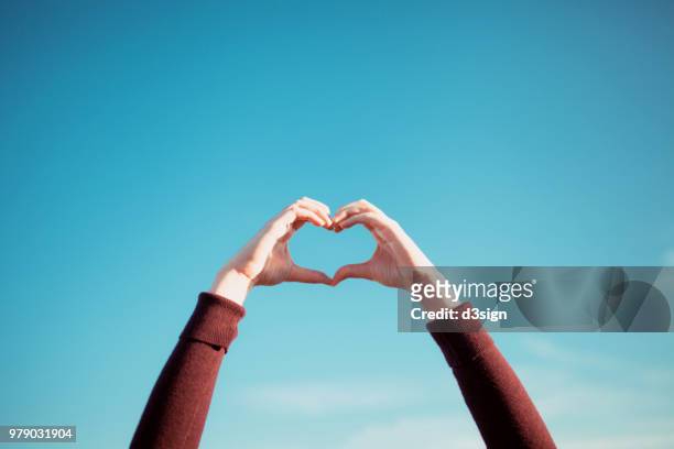 woman's hand gesturing a heart shape over clear blue sky and warm sunlight - hands happy ストックフォトと画像