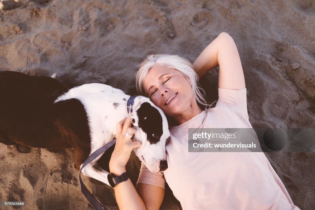 Lächelnde ältere Frau mit Hund am Strand liegend