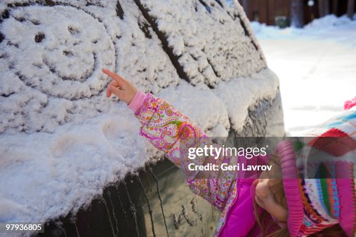 Girl making a happy face shape on a car with snow