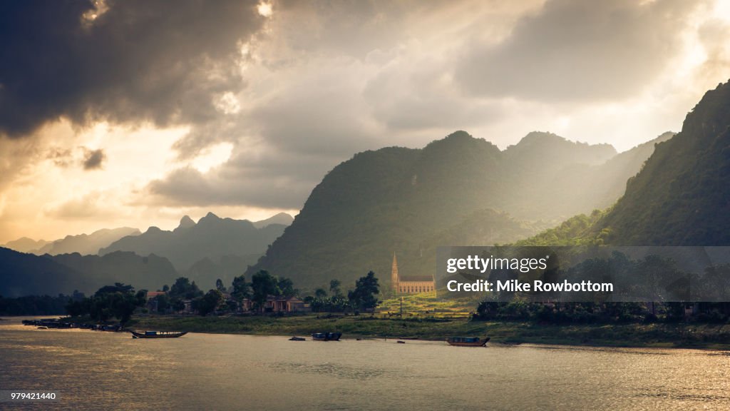 Church and forest covered mountains on riverbank at sunset under cloudy sky, Quang Binh Province, Vietnam