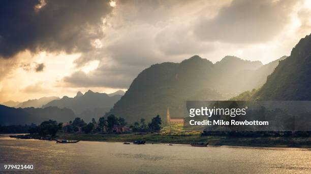 church and forest covered mountains on riverbank at sunset under cloudy sky, quang binh province, vietnam - phong nha kẻ bàng national park stock pictures, royalty-free photos & images