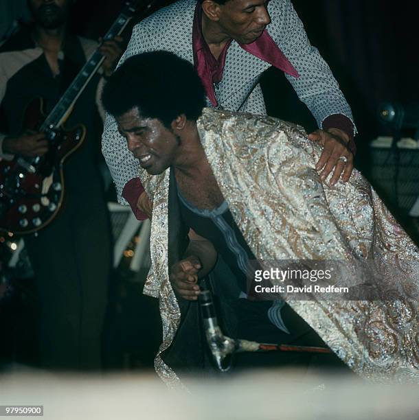 American soul singer and songwriter James Brown has a cape placed over him during a performance on stage with the J.B.'s, including guitarist Catfish...